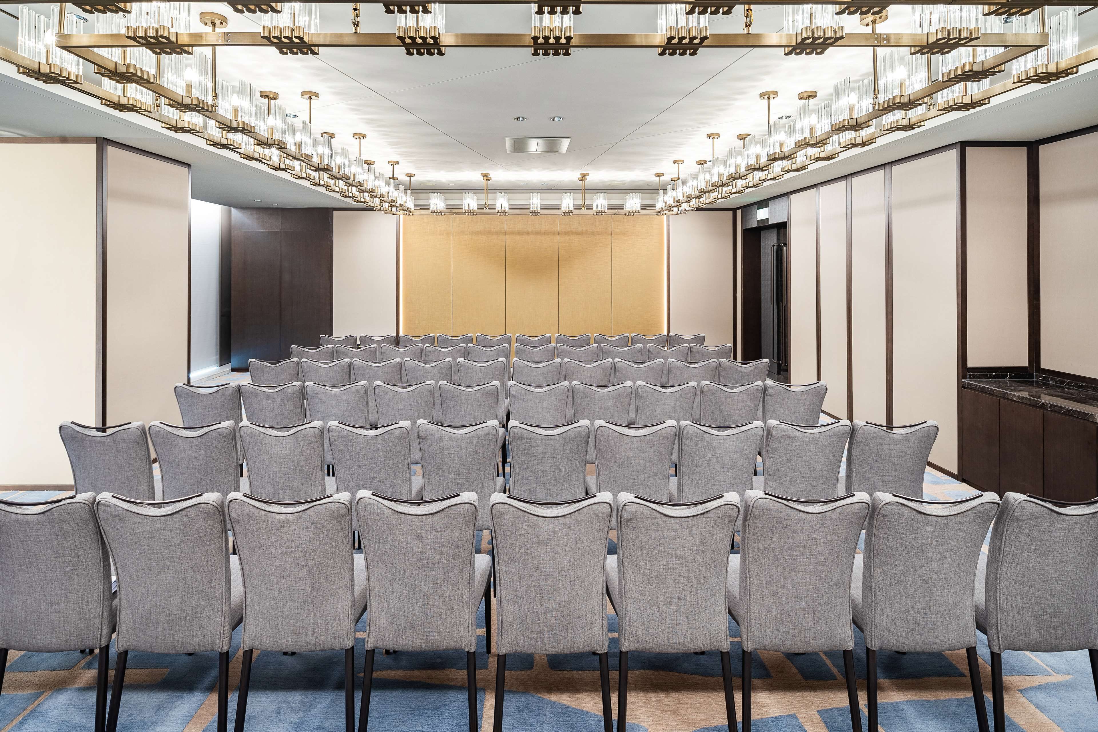 Una sala de conferències amb files de cadires grises, una pantalla gran i un llum modern.