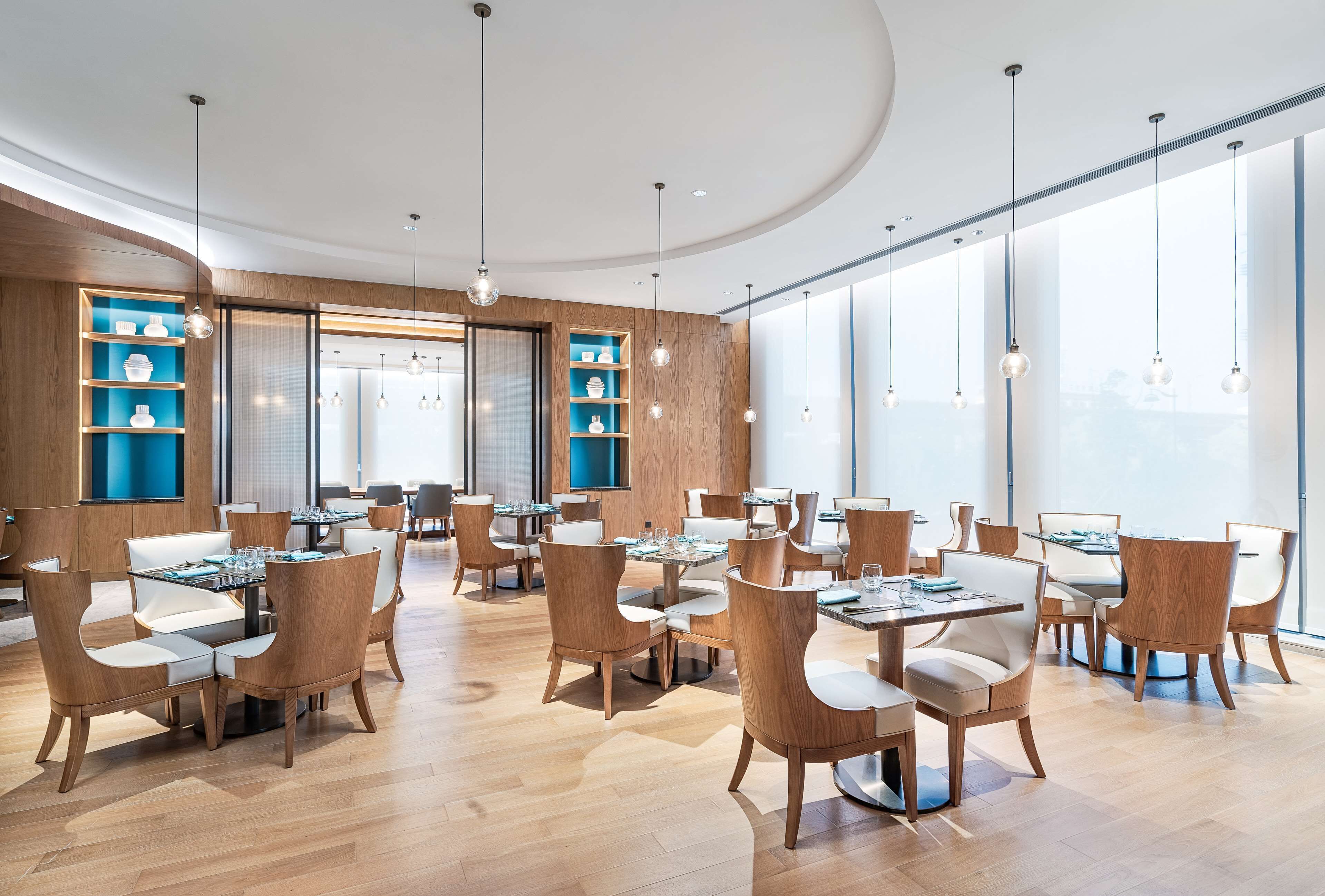 A modern dining area with wooden furniture and glass tables, large windows, and pendant lighting.