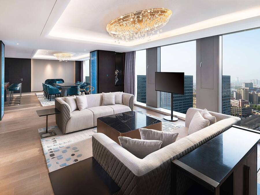 Une chambre d’hôtel moderne dotée d’une grande fenêtre, d’un lustre, d’un canapé, d’une table basse et d’un téléviseur à écran plat.