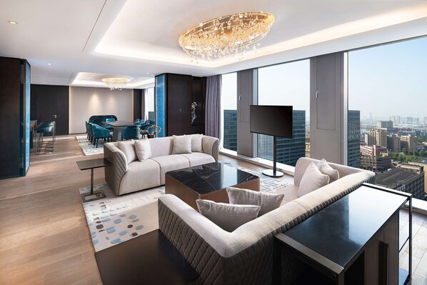 A modern hotel room with a large window, a chandelier, a sofa, a coffee table, and a flat-screen TV.