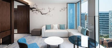 A modern living room with a sofa, a round coffee table, and a glass table. There is a large window with a view of buildings.