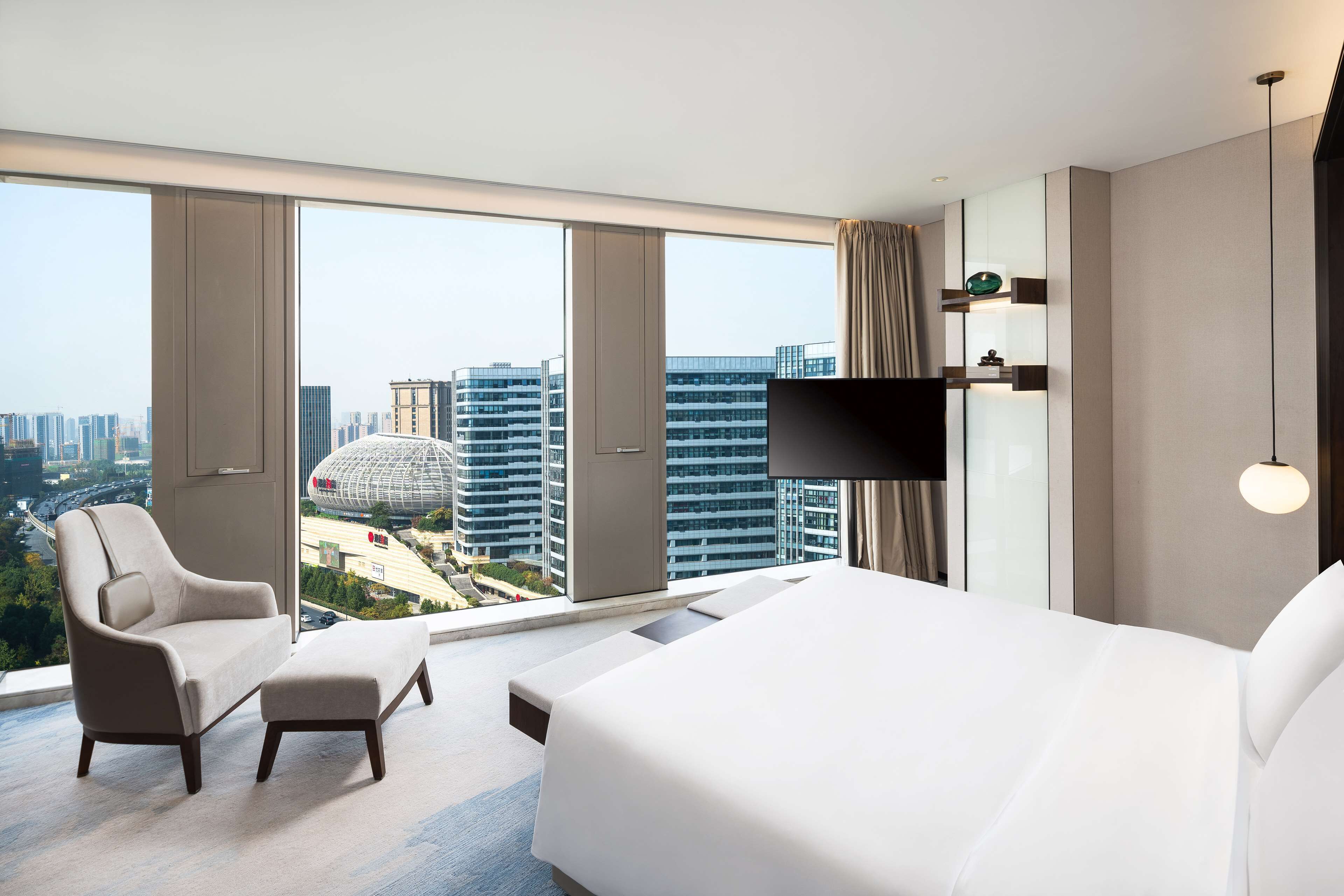 A modern hotel room with a large bed, a sofa, and a chair. It features a city view through large windows.