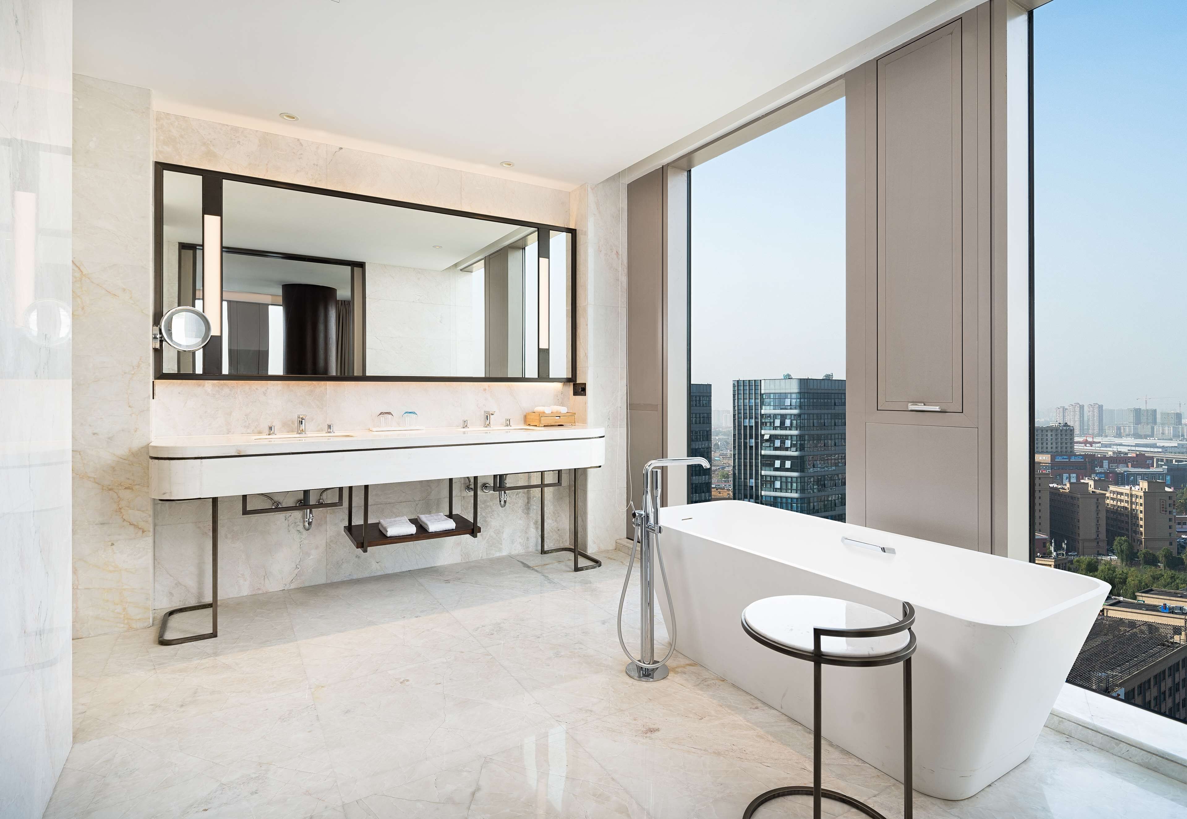 A modern bathroom with a large mirror, dual sinks, a bathtub, and a city view.