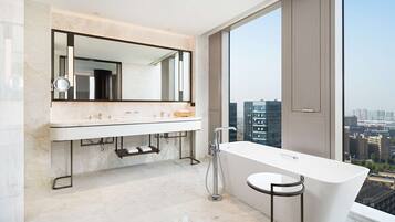 A modern bathroom with a large mirror, dual sinks, a bathtub, and a city view.