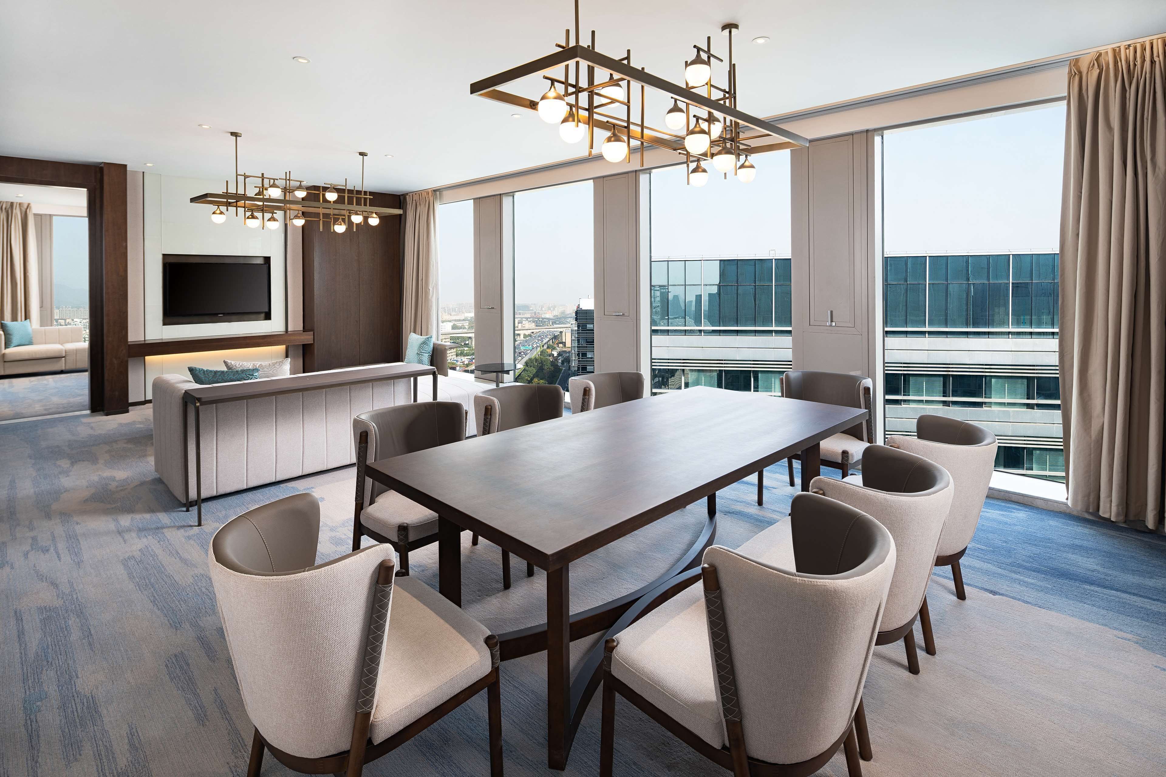 A modern hotel room with a large dining table, comfortable seating, a TV mounted on the wall, and a city view through large windows.