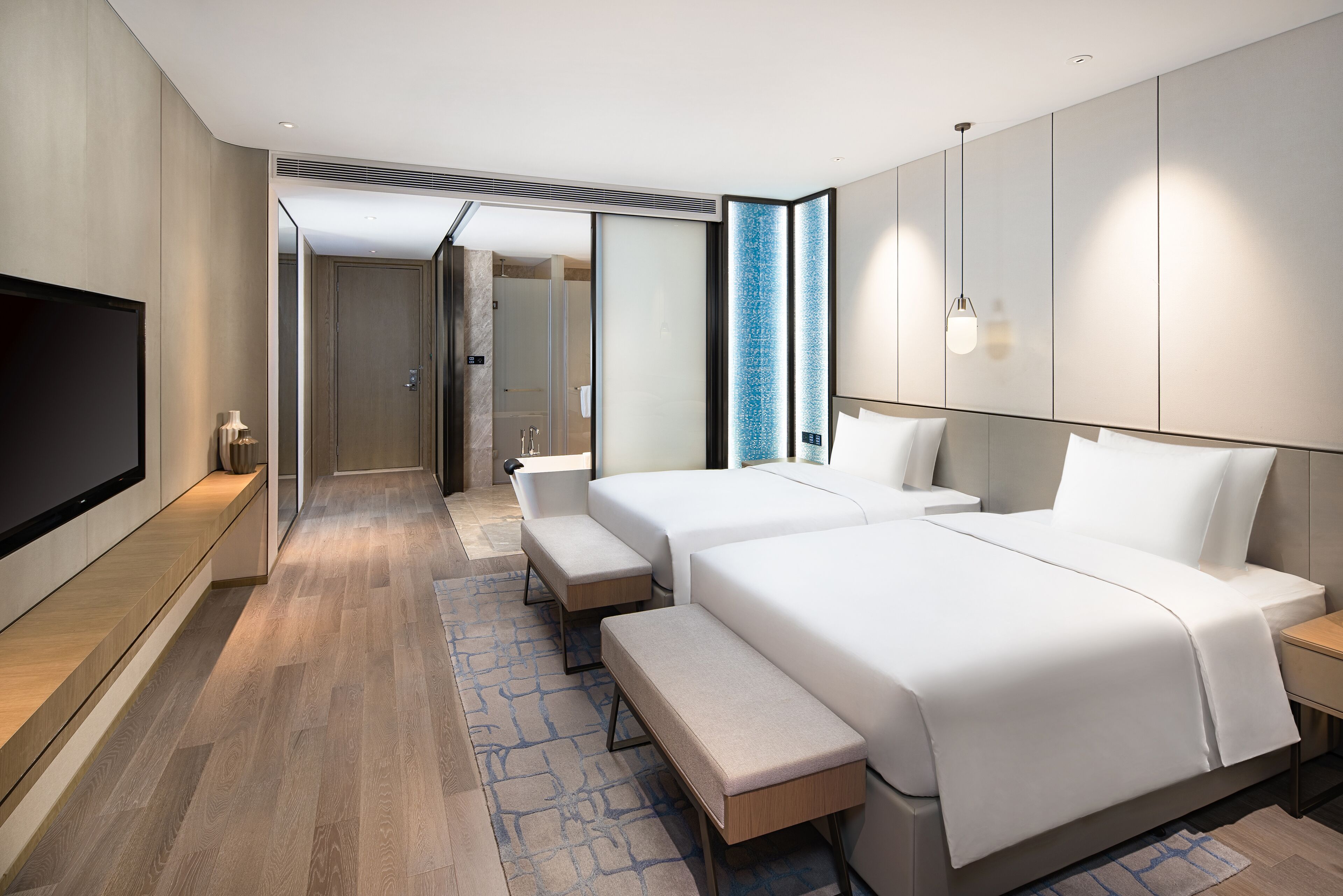 Una habitación de hotel moderna con dos camas, un banco, un televisor y un baño visible a través de una puerta abierta.