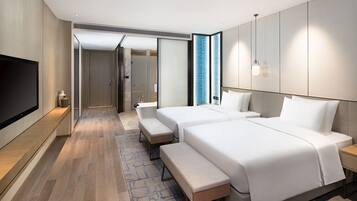A modern hotel room with two beds, a bench, a TV, and a bathroom visible through an open door.