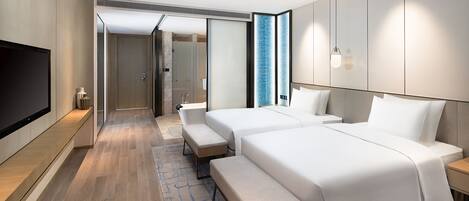 Una habitación de hotel moderna con dos camas, un banco, un televisor y un baño visible a través de una puerta abierta.