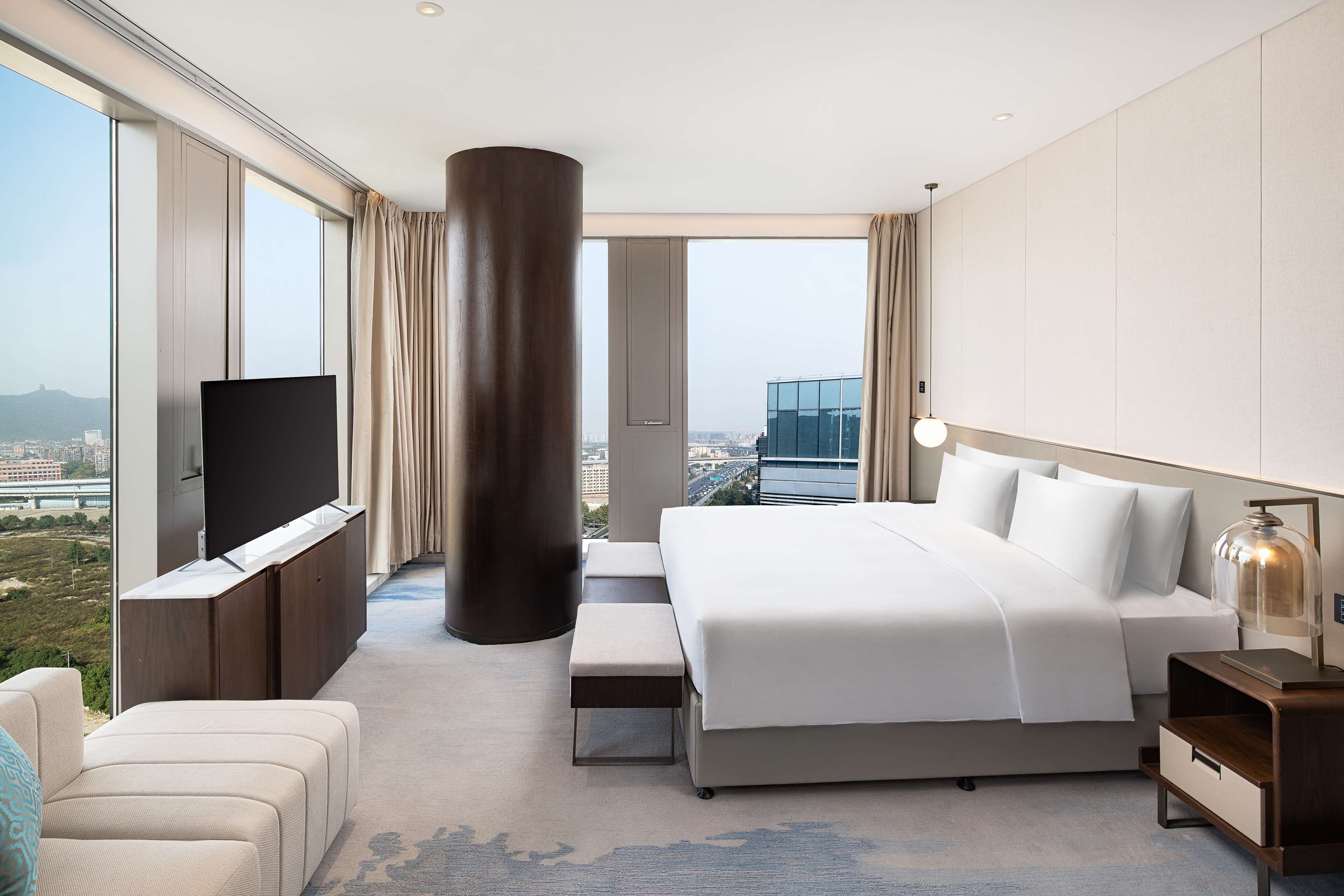 A modern hotel room with a large bed, a TV, a bench, and a view of the city.