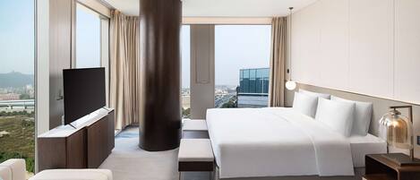 A modern hotel room with a large bed, a TV, a bench, and a view of the city.