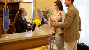 Un comptoir d’accueil avec un réceptionniste, un client tenant une fleur et un homme avec une valise.