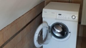 A front-loading washing machine in a corner of a room with tiled flooring and a sloped ceiling.