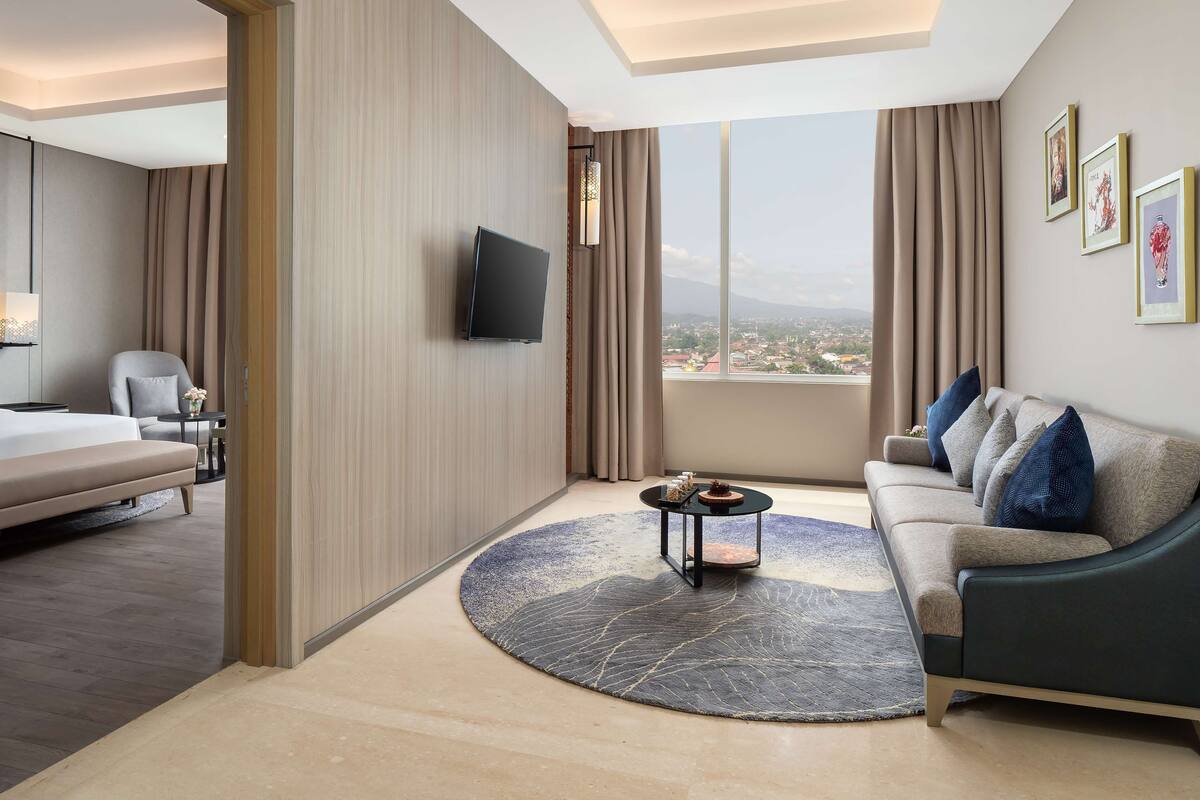 A modern hotel room with a bed, a sofa, a TV, and a large window offering a city view.