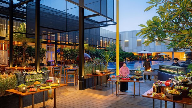 An outdoor dining area with wooden tables, a pool, and a variety of plants and trees. - Radisson Lampung Kedaton (Bandar Lampung)
