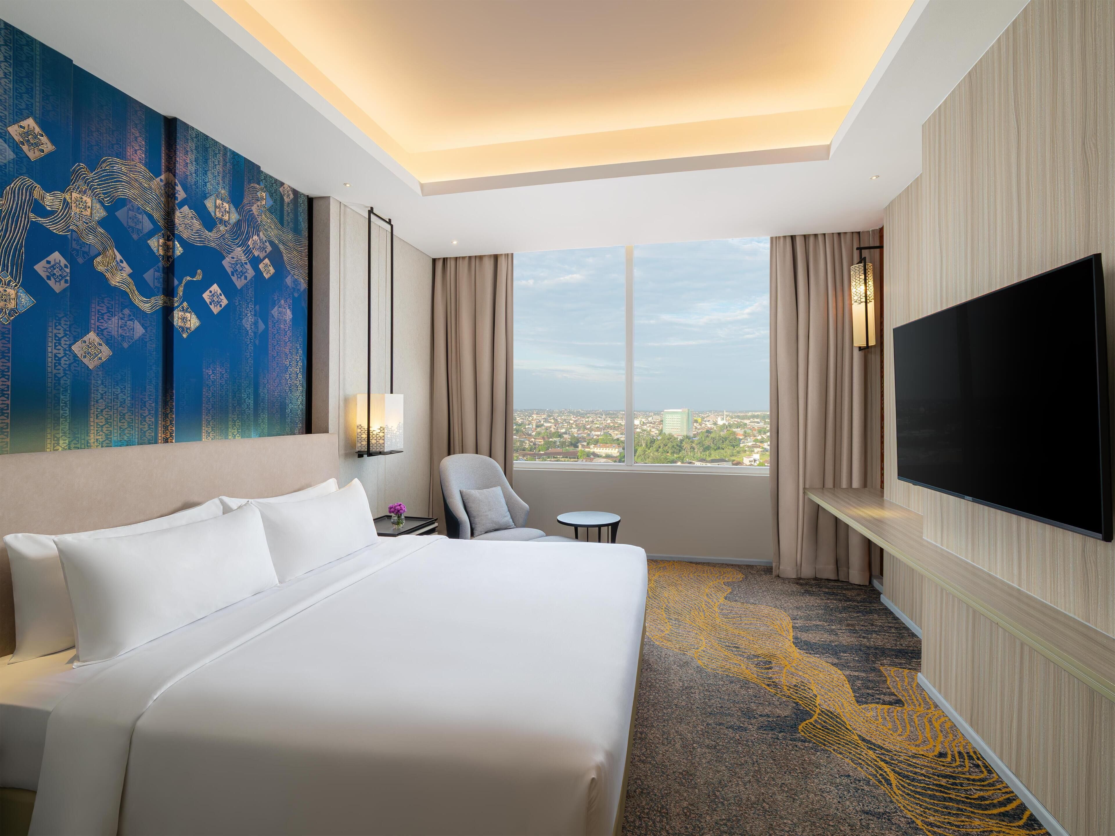A hotel room with a large bed, a chair, a small table, a TV, and a city view through the window.