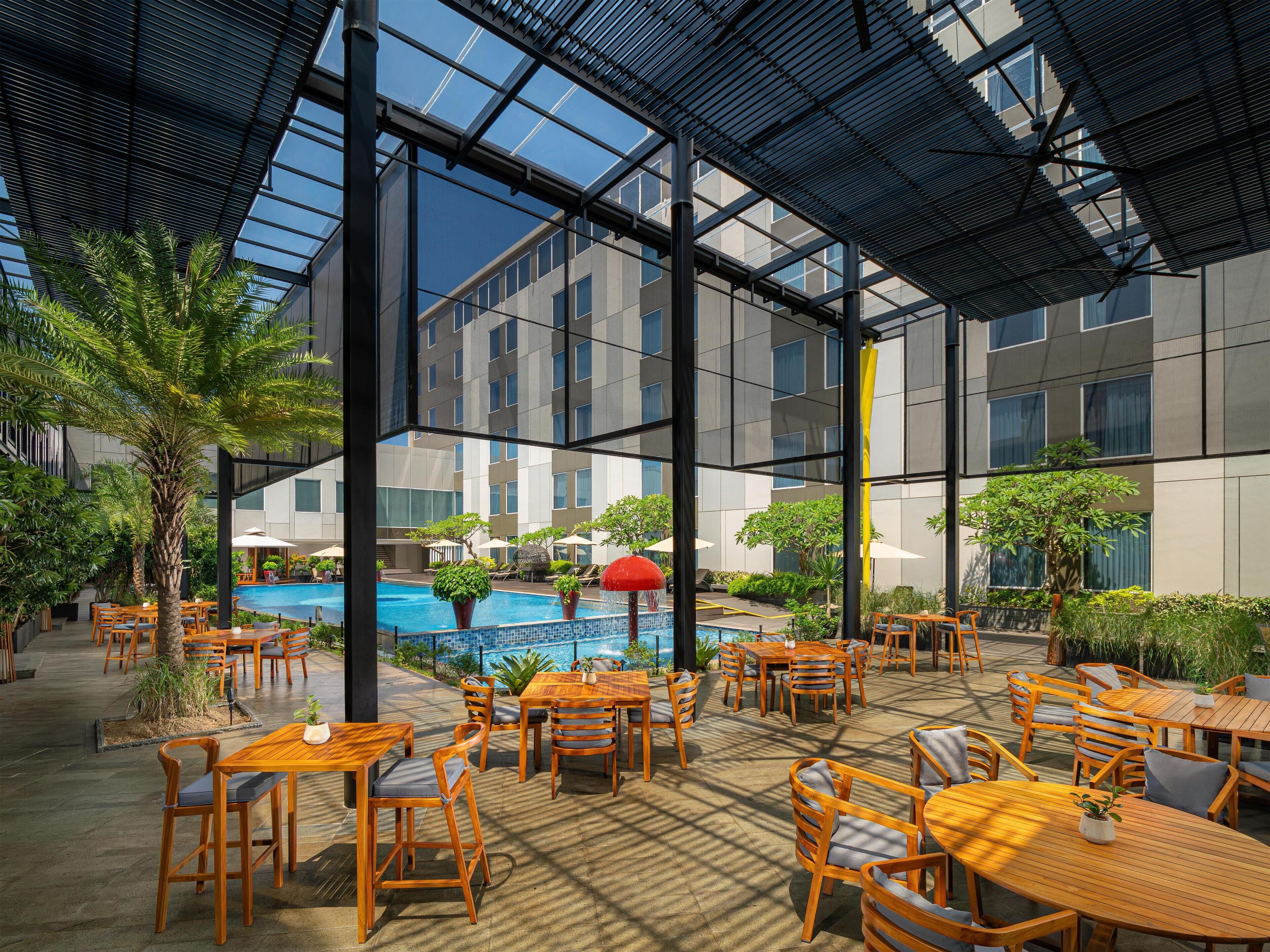 An indoor pool area with wooden tables and chairs, surrounded by greenery and modern architecture.