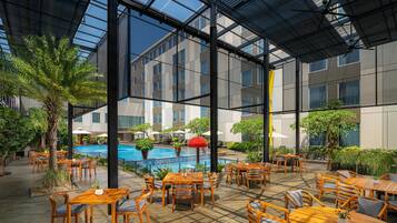 An indoor pool area with wooden tables and chairs, surrounded by greenery and modern architecture.
