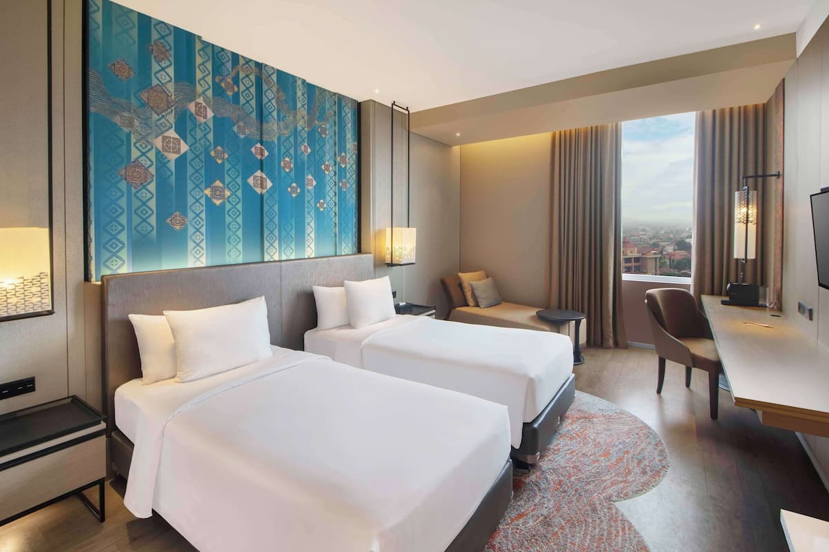 A hotel room with two beds, a desk, a chair, a TV, and a large window with curtains.