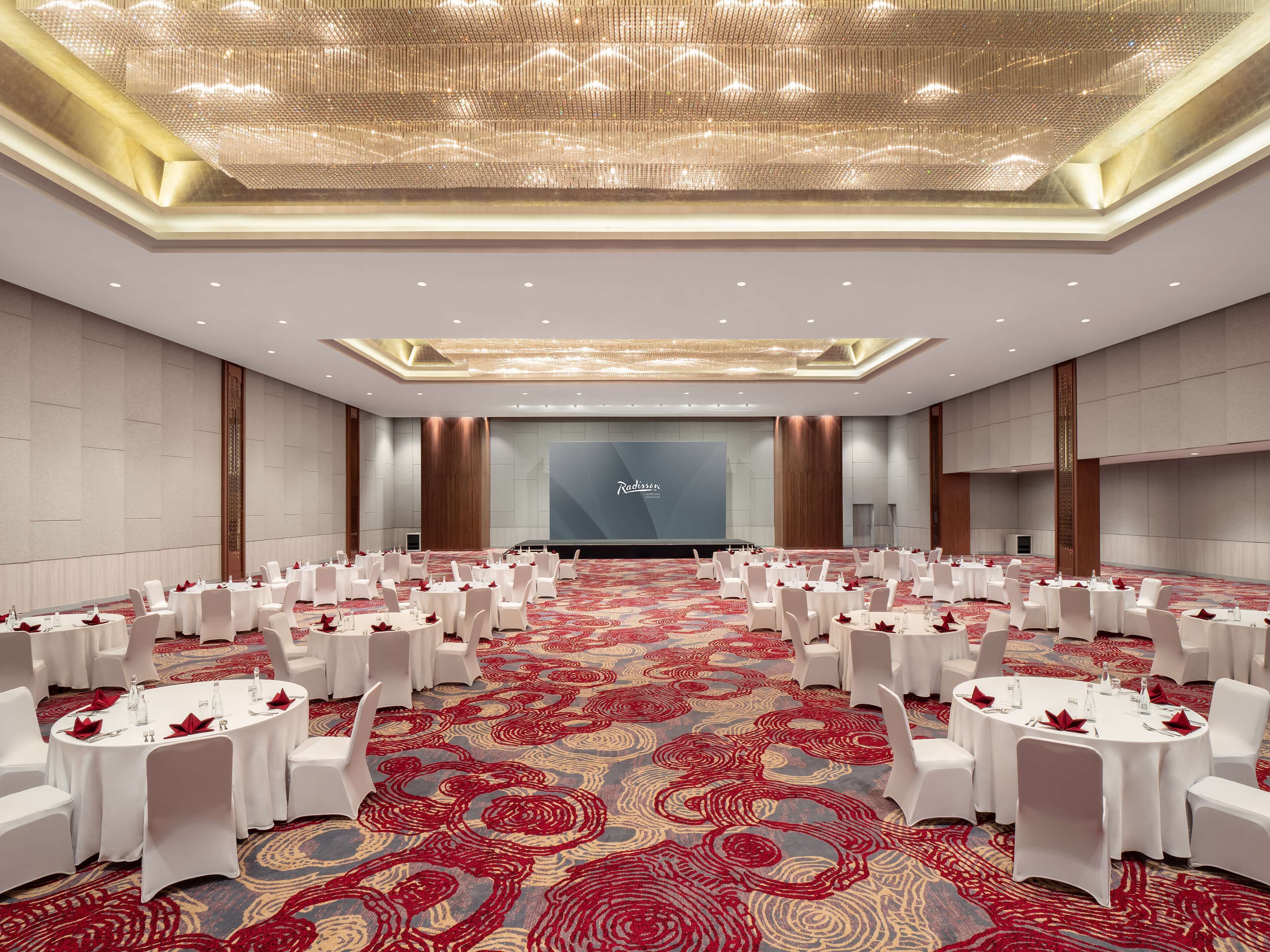 Une grande salle de conférence avec des tables rondes et des chaises blanches, un tapis rouge et un grand écran à l’avant.