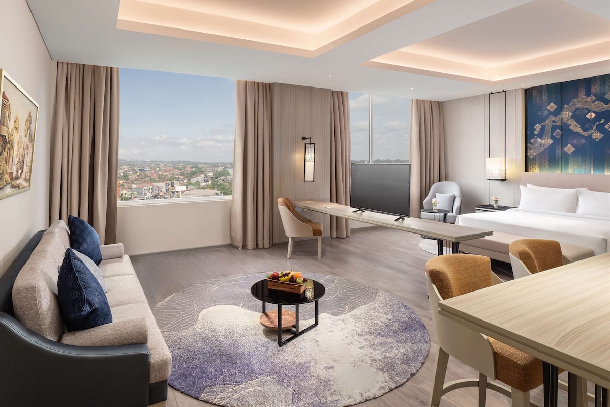 A modern hotel room with a large bed, a sofa, a dining table, and a view of the city.