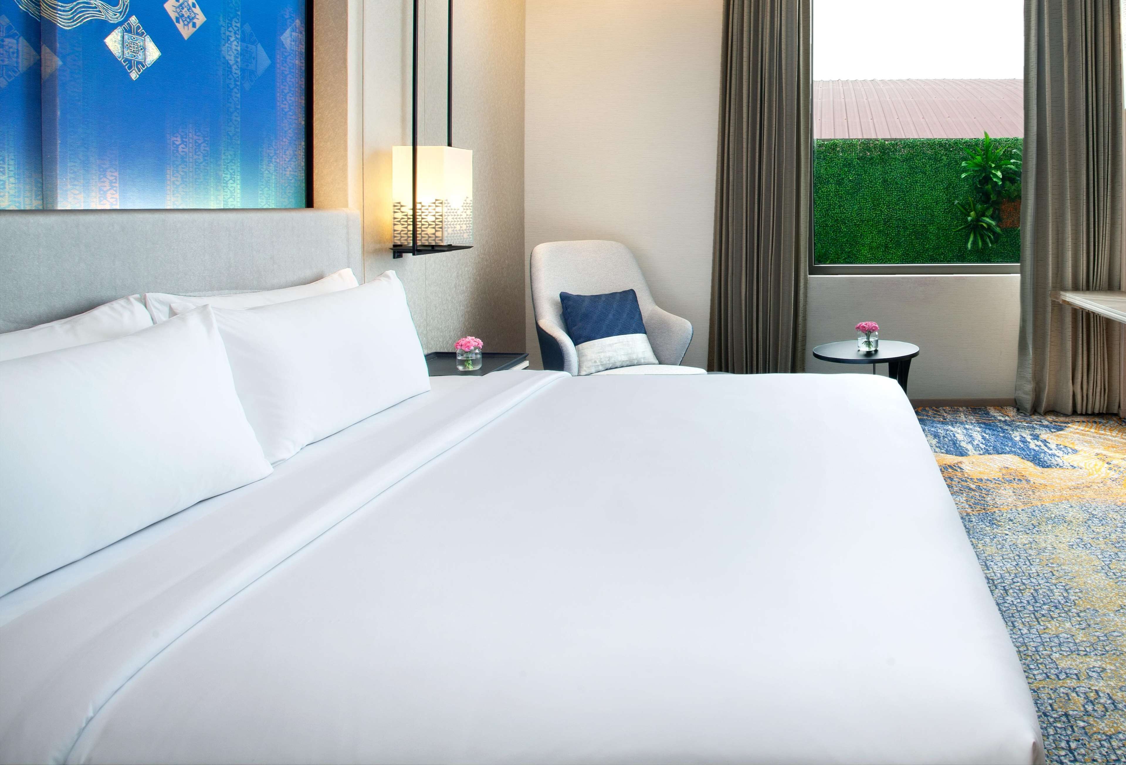 Ein Hotelzimmer mit einem großen Bett, einem Sessel, einem kleinen Tisch mit einer Vase und Blick auf einen grünen Heckenstreifen durch ein Fenster.