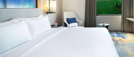 Ein Hotelzimmer mit einem großen Bett, einem Sessel, einem kleinen Tisch mit einer Vase und Blick auf einen grünen Heckenstreifen durch ein Fenster.