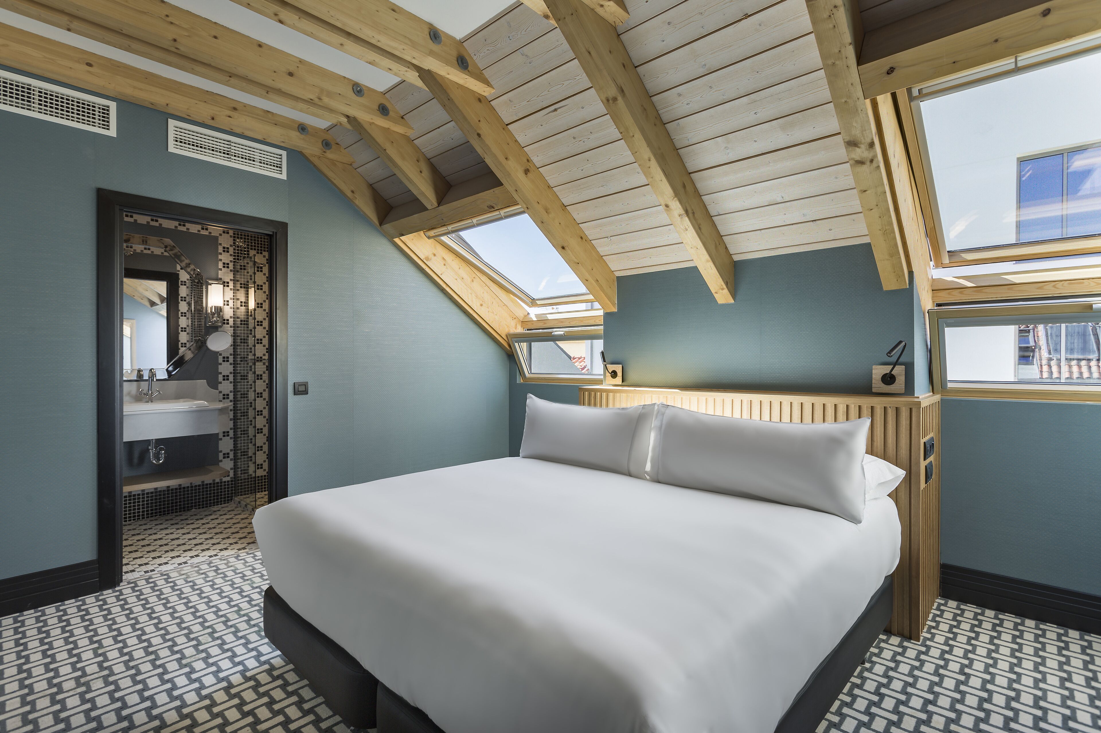 Ein Schlafzimmer mit einem großen Bett, einem Oberlicht, Holzbalken und einem Badezimmer mit Waschbecken und Spiegel.
