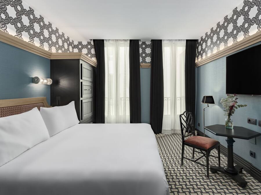 Una habitación de hotel con una cama grande, una mesita con un florero, una silla y un televisor en la pared.