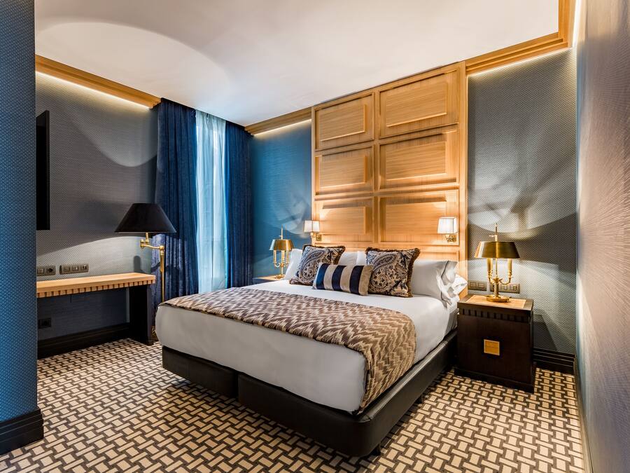 A modern hotel room with a large bed, two bedside tables with lamps, a built-in headboard, and a patterned floor.