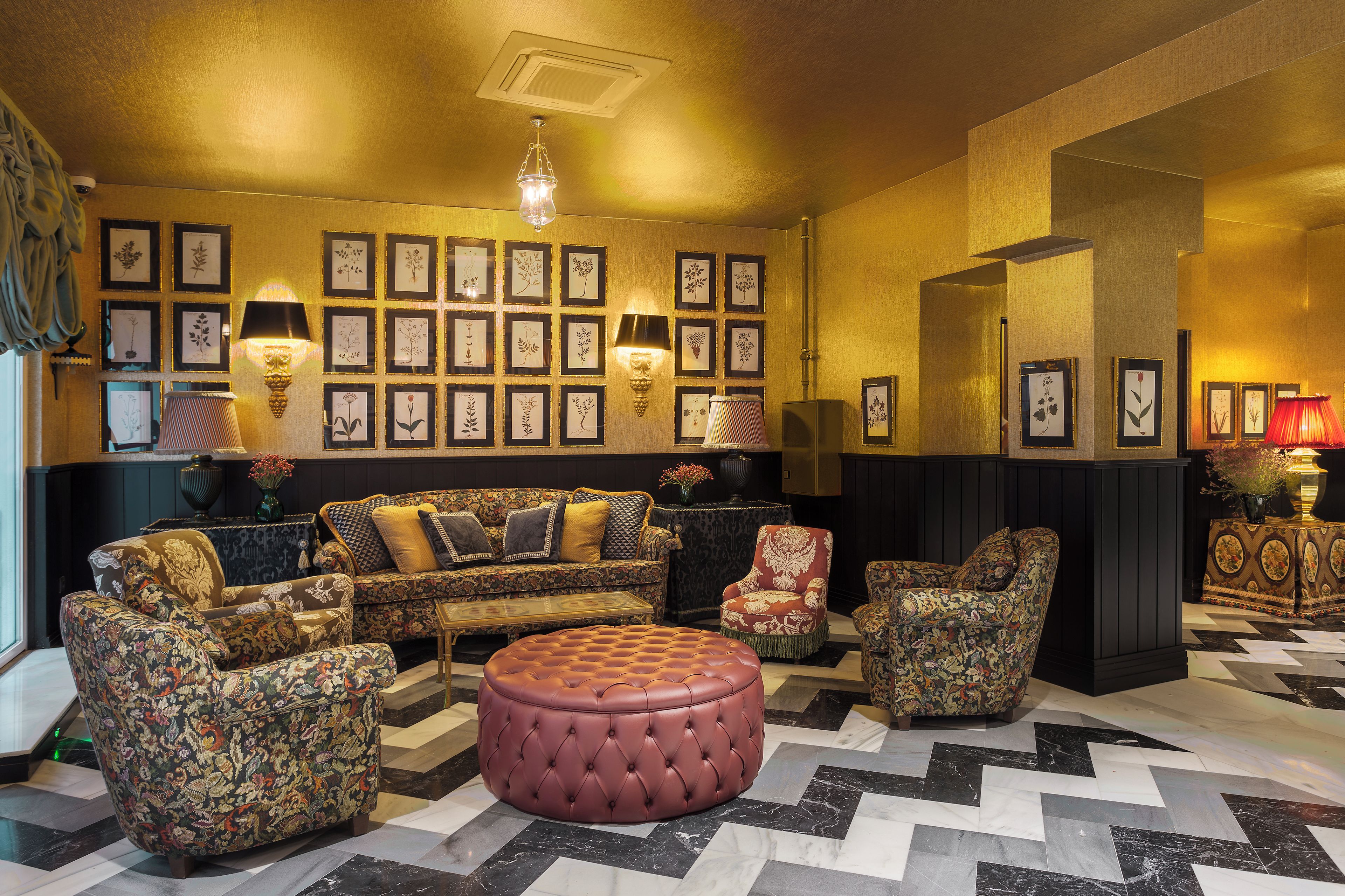 Une pièce comprenant un canapé à motifs, des fauteuils, un pouf capitonné et un mur décoré d’illustrations botaniques encadrées.