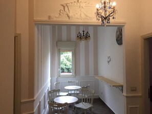 Un comedor con ventana, sillas blancas, una araña y una pared blanca con un reloj.