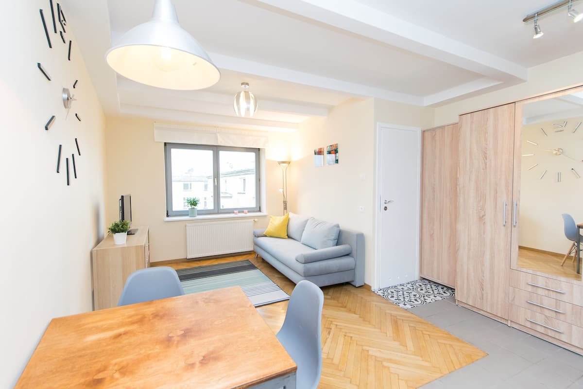 A modern living room with a wooden dining table, a grey sofa, a large window, and a wardrobe.
