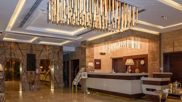 Zona de recepción de un hotel con pared de mármol, mostrador de recepción, una araña y una escalera.
