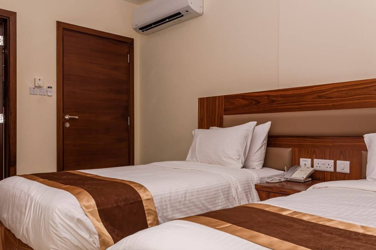 Habitación de hotel con dos camas, cabeceros de madera, ropa de cama blanca, teléfono en la mesita de noche y un equipo de aire acondicionado.