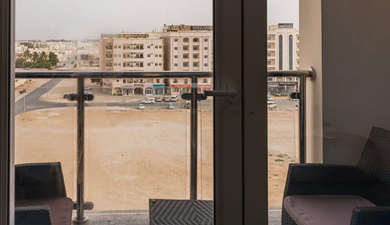 A view from a balcony with a metal railing, overlooking a street with buildings and a roundabout.