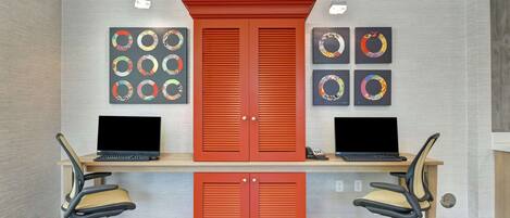 A workspace with two desks, each with a laptop and an office chair. There are two red cabinets and artwork on the wall.