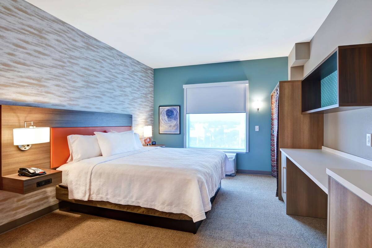 Ein Hotelzimmer mit Bett, Nachttisch, Lampe und einem Fenster mit Jalousien.