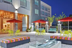 An outdoor patio area with yellow chairs and red umbrellas, a glass-top table, and a stone wall. - Home2 Suites by Hilton Charlotte Piper Glen (Charlotte)