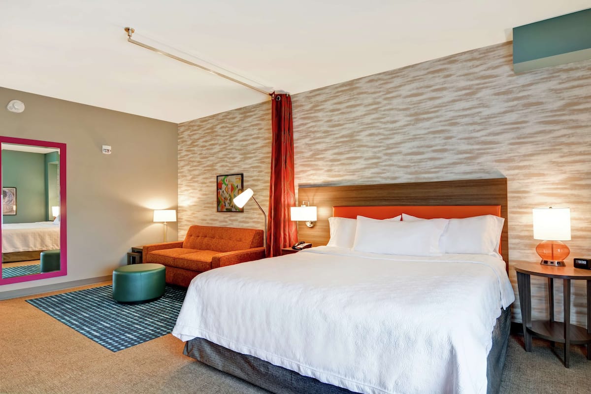 Ein Hotelzimmer mit einem großen Bett, einem Schreibtisch mit Lampe, einem Sessel, einem kleinen Hocker und einem Spiegel mit rosafarbenem Rahmen.
