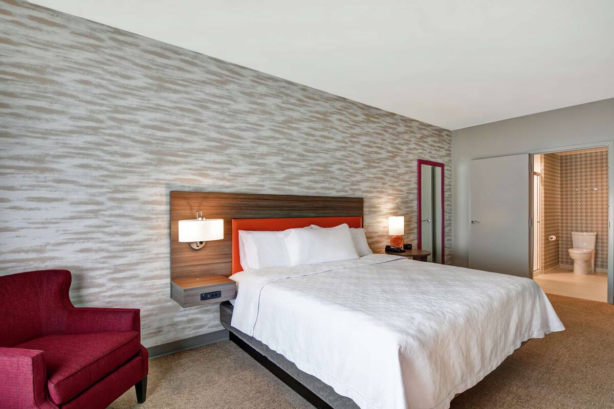 Ein Hotelzimmer mit einem Bett, einem roten Sessel, einem Nachttisch und einem durch eine offene Tür einsehbaren Badezimmer.