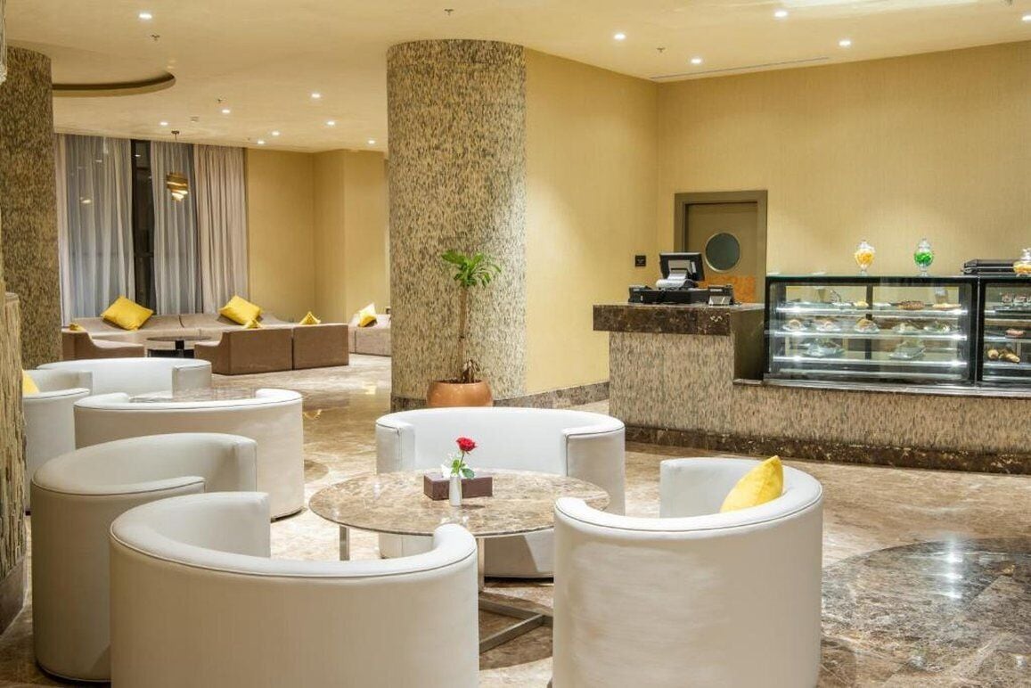 En modern entréhall med en sittgrupp bestående av vita soffor och ett soffbord i marmor. En stor pelare med en växt, en receptionsdisk och ett vitrinskåp med matvaror syns också.