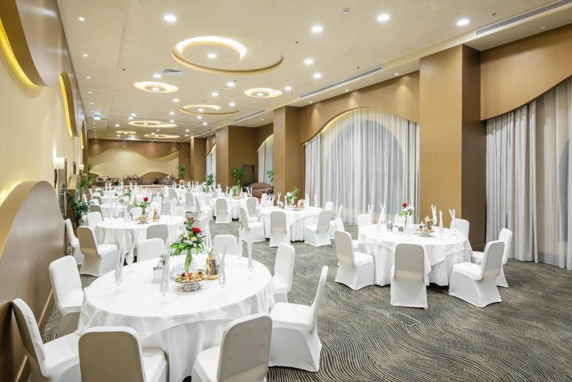 Une salle de banquet avec des tables rondes disposées pour des événements, recouverte de nappes blanches, dotée de chaises et d’un sol moquetté.