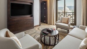 Habitación de hotel con una amplia televisión de pantalla plana en la pared, un conjunto de sofá, una mesa de centro redonda y vistas a un paisaje urbano con edificios de cúpulas.