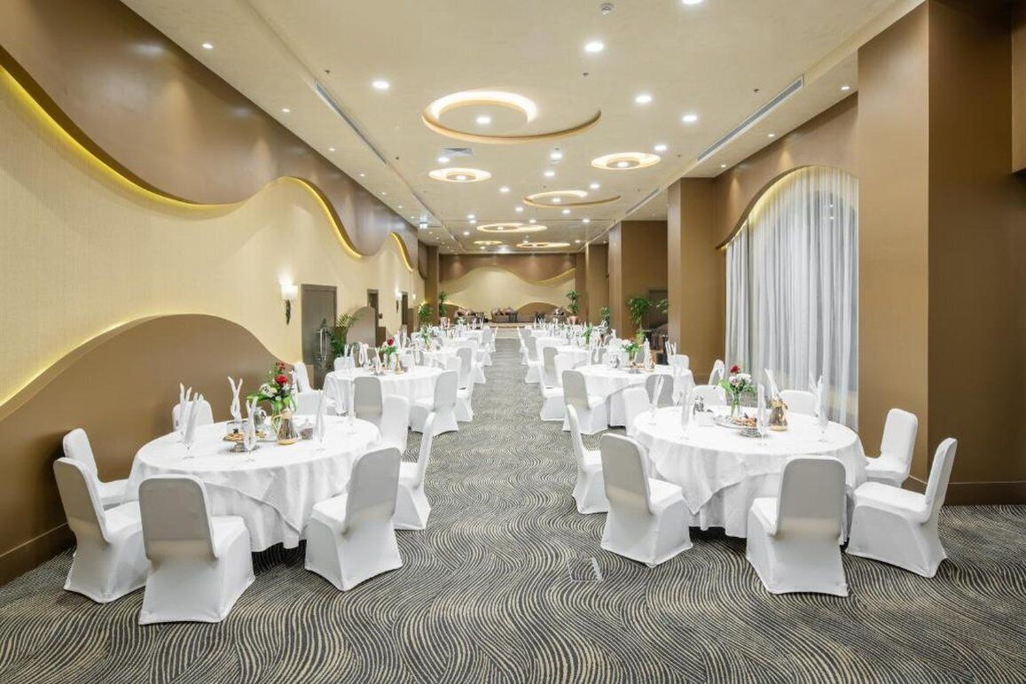 Une salle de banquet avec des tables rondes dressées pour le repas, des chaises blanches et un sol recouvert de moquette.