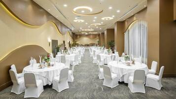 Une salle de banquet avec des tables rondes dressées pour le repas, des chaises blanches et un sol recouvert de moquette.