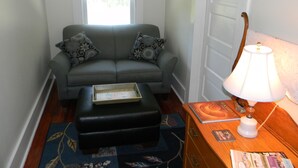 Una sala de estar con un sofá gris, cojines estampados, un reposapiés negro, una mesita de madera con una lámpara y una ventana con cortina.