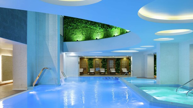 A modern indoor swimming pool with a clear view of the pool area, a seating area with chairs, and a decorative wall with greenery.