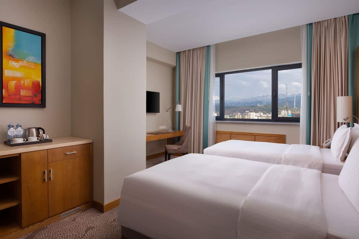 Ett hotellrum med två sängar, ett skrivbord, en stol, en TV och ett fönster med utsikt över staden.