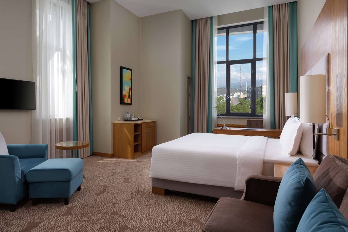 Ett hotellrum med en säng, ett skrivbord, en stol, en TV och ett fönster med utsikt över staden.