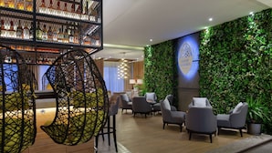 A modern lounge area with a wall-mounted bar displaying various liquor bottles, seating with a geometric hanging chair, and a wall with a vertical garden labeled "ROOF GARDEN".